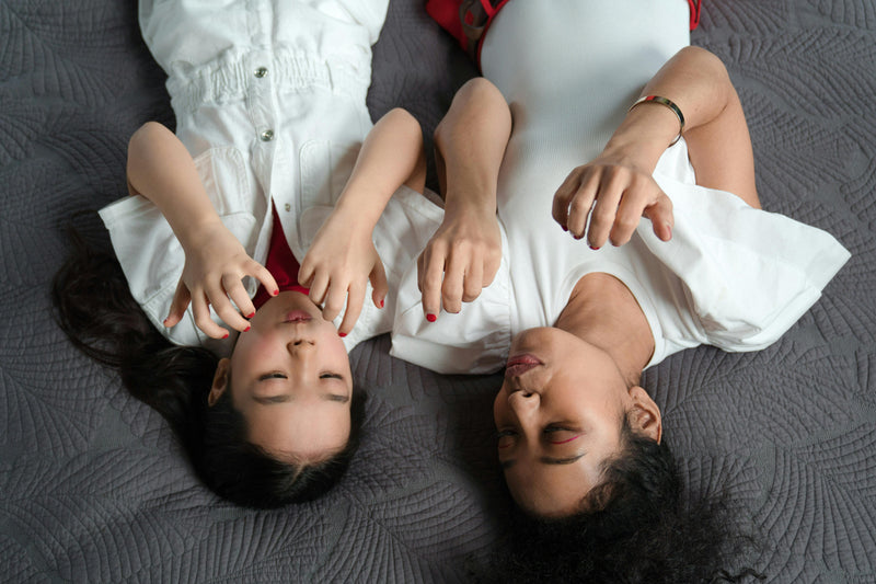 Mother and daughter lying together in coordinated white outfits, sharing a quiet, tender moment that reflects the beauty of harmonised family style.